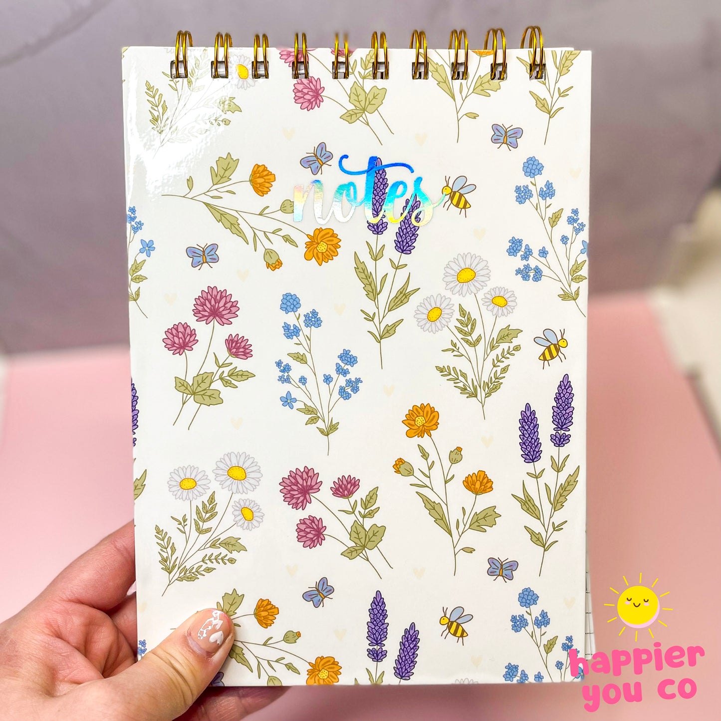 Notebook with floral pattern held by a hand, featuring the brand 'Happier You Co'.