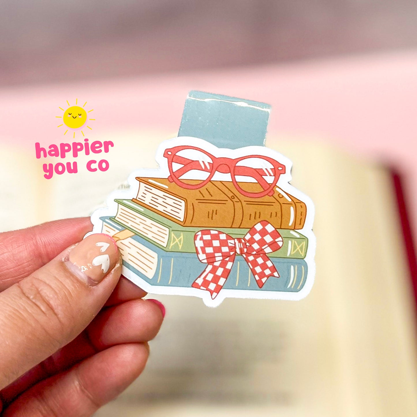 Magnetic bookmark with books and glasses held by a hand, featuring 'happier you co' text.