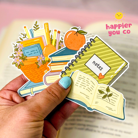 A hand with light blue nail polish holds a cluster of colorful planner and study-themed stickers shaped like books, notebooks, flowers, pencils, and an open book. The background is a softly blurred open book with pink lighting, and the text 'happier you co' appears in the top right.