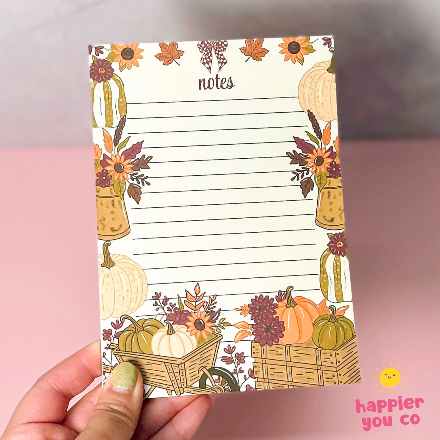 A hand holding a vertical lined notepad with an autumn harvest border featuring pumpkins, sunflowers, leaves, and baskets. The notepad has the word 'notes' at the top and a pink 'happier you co' logo in the bottom right corner of the photo.