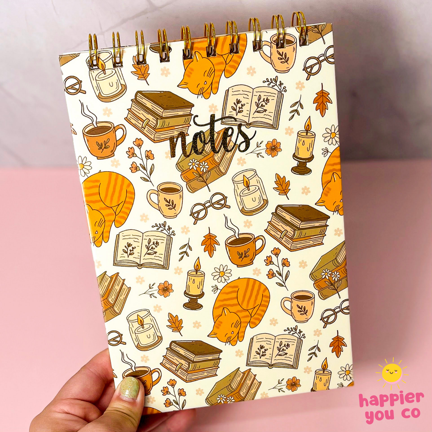 A hand holds a spiral-bound notebook with a cozy autumn-themed illustration pattern featuring orange cats, books, coffee cups, candles, and leaves on a cream background. The brand name 'happier you co' is visible in the bottom right corner.