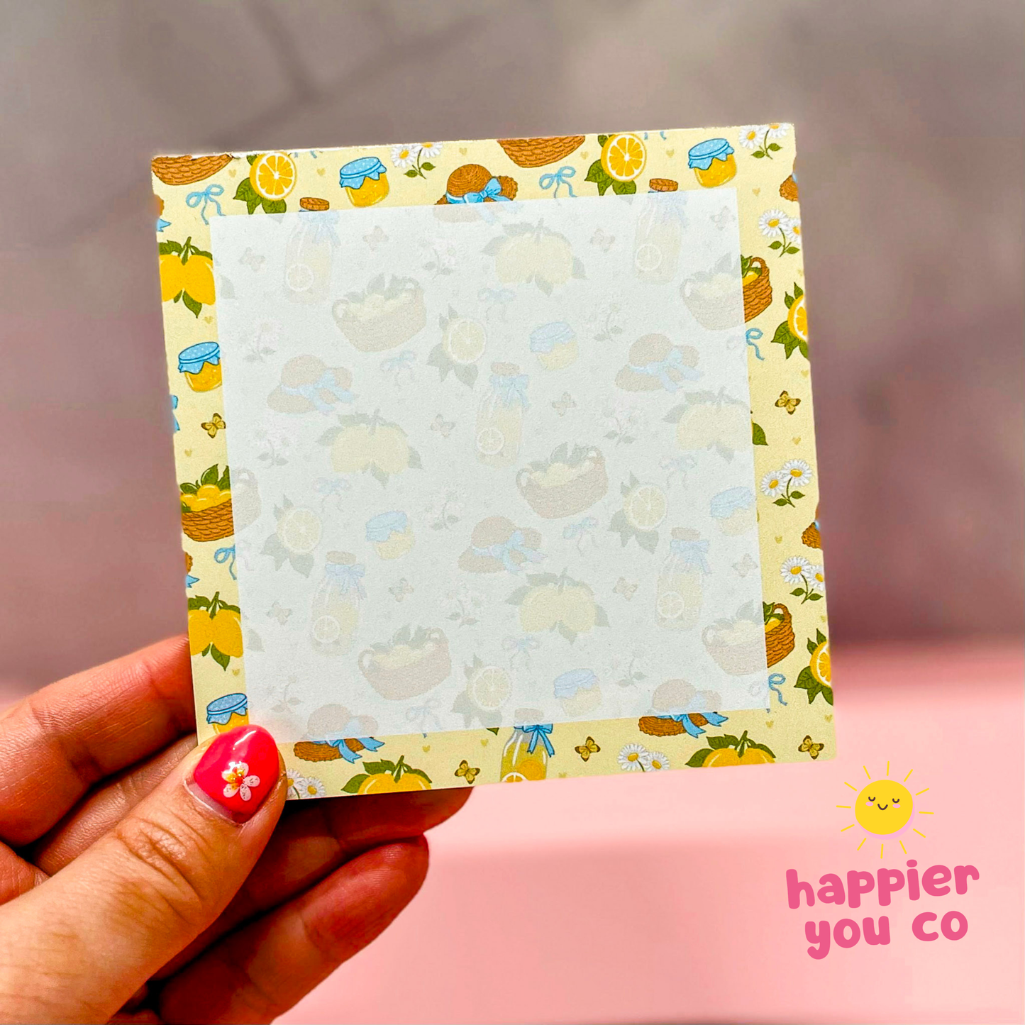Hand holding a decorative card with a colorful border featuring lemons and birds, on a pink background with 'happier you co' branding.