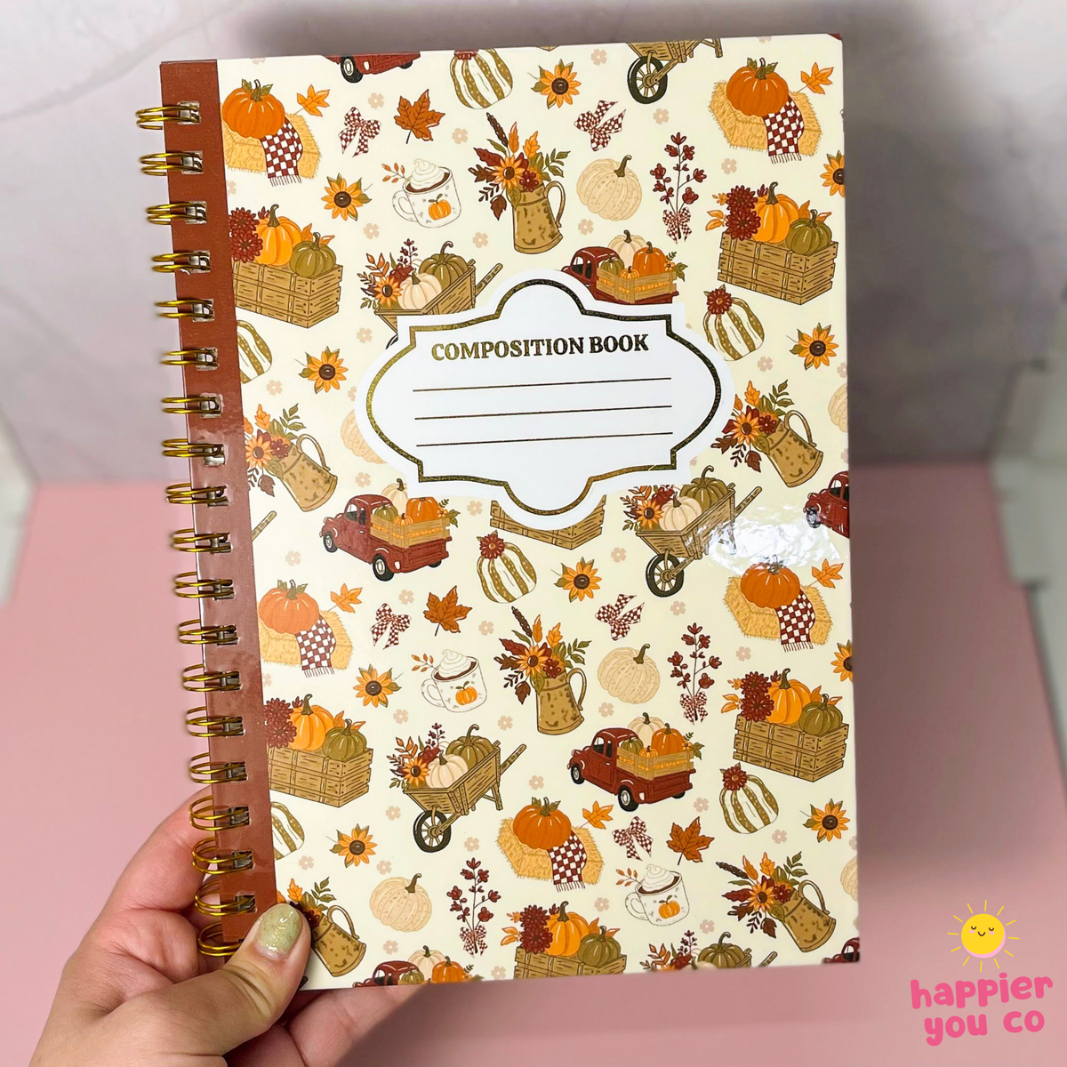 A hand holds a spiral-bound composition notebook with a fall harvest pattern featuring pumpkins, autumn leaves, baskets, and a central label that reads 'COMPOSITION BOOK'. The background is a soft pink surface with a light gray wall behind it.