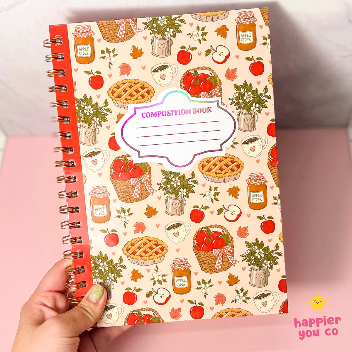 A hand holds a spiral-bound composition book with a cream-colored cover decorated with autumn-themed illustrations of pies, apples, strawberries, and coffee cups. The book has a red-orange spiral binding and a white label on the front that reads 'COMPOSITION BOOK'.