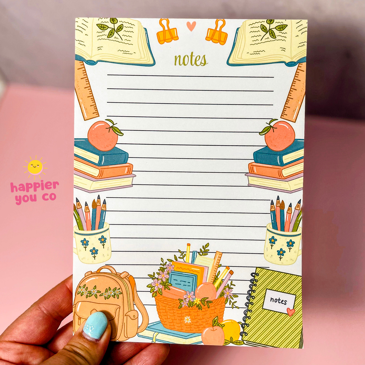 A hand with light blue nail polish holds a decorative lined notepad. The notepad features school-themed illustrations including books, pencils, an apple, a backpack, and a basket, with the word 'notes' at the top.