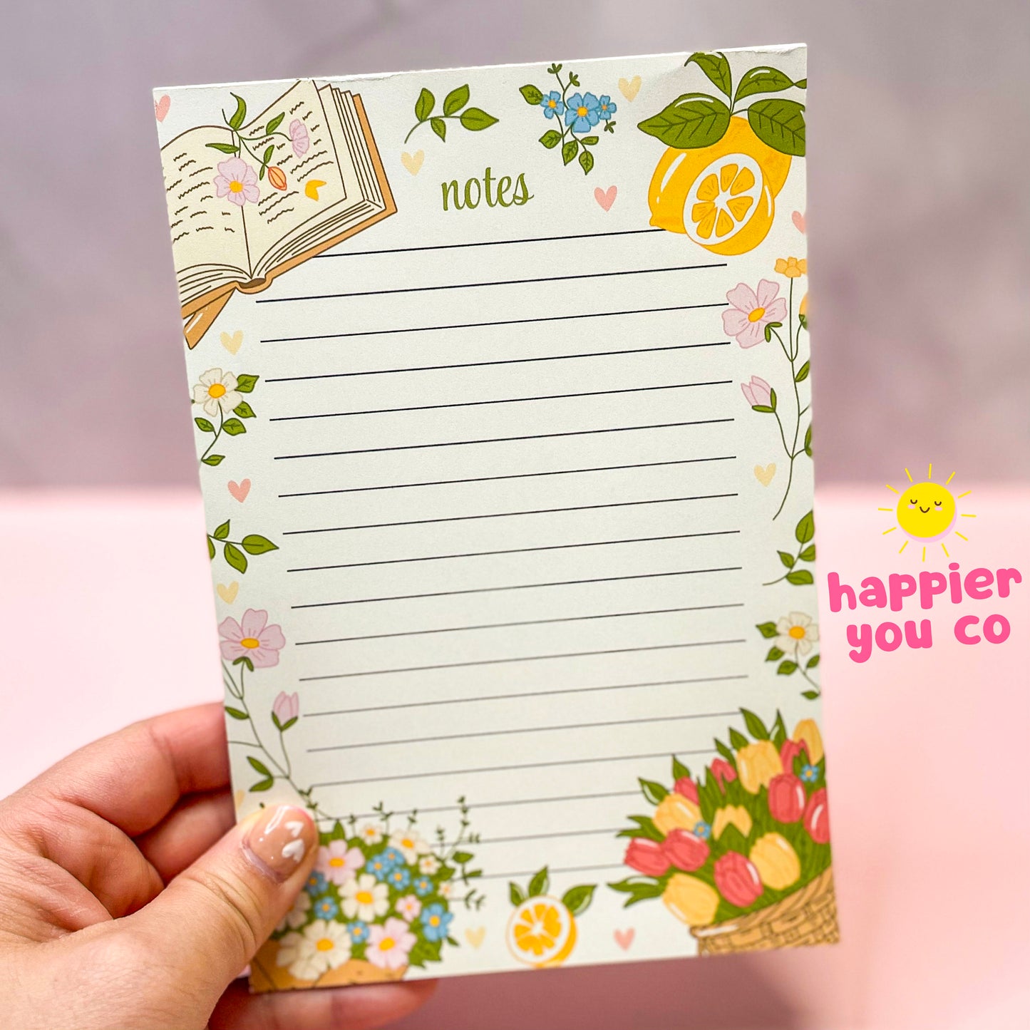 Notepad with floral and lemon design held by a hand, branded 'happier you co'.