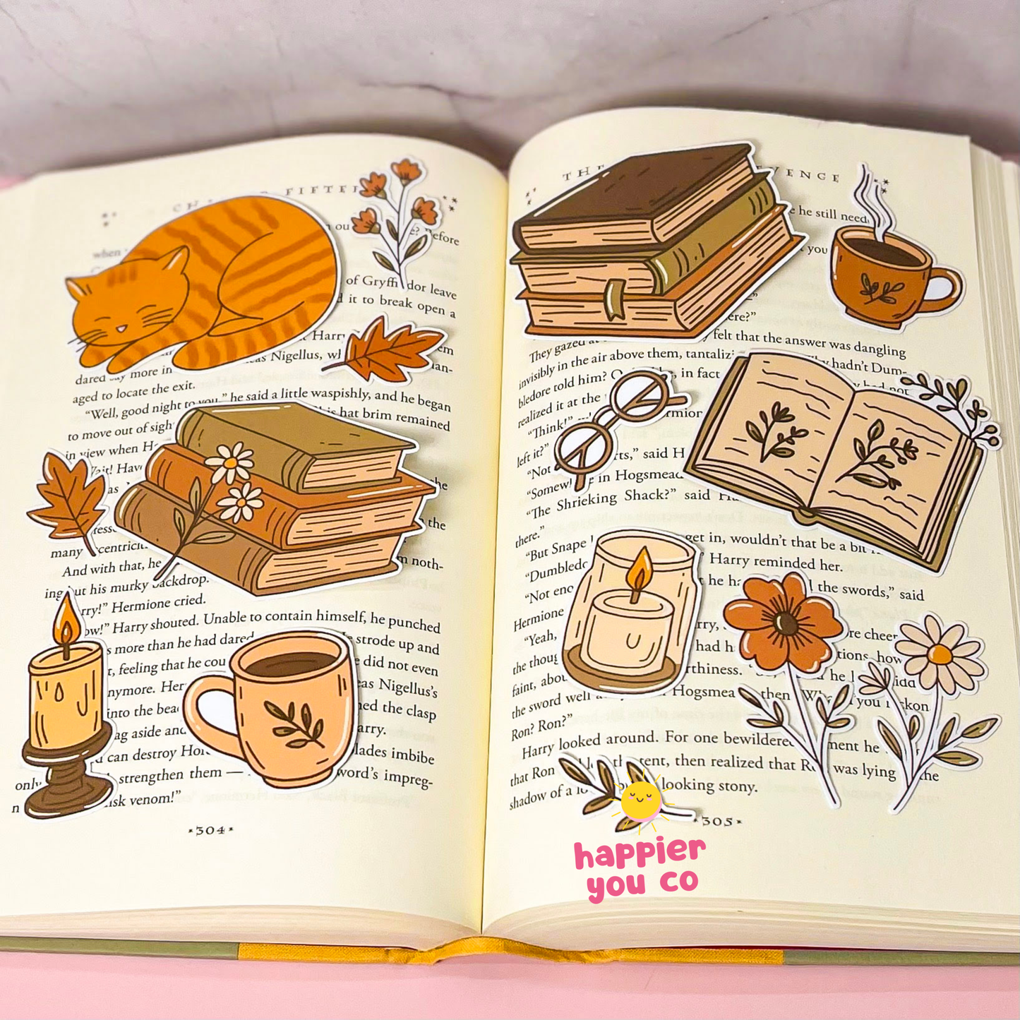An open decorative book with illustrated autumn and cozy themes, including an orange cat, stacked books, a mug, candles, flowers, and small drawings layered over printed text. The phrase 'happier you co' appears in pink at the bottom of the right page.