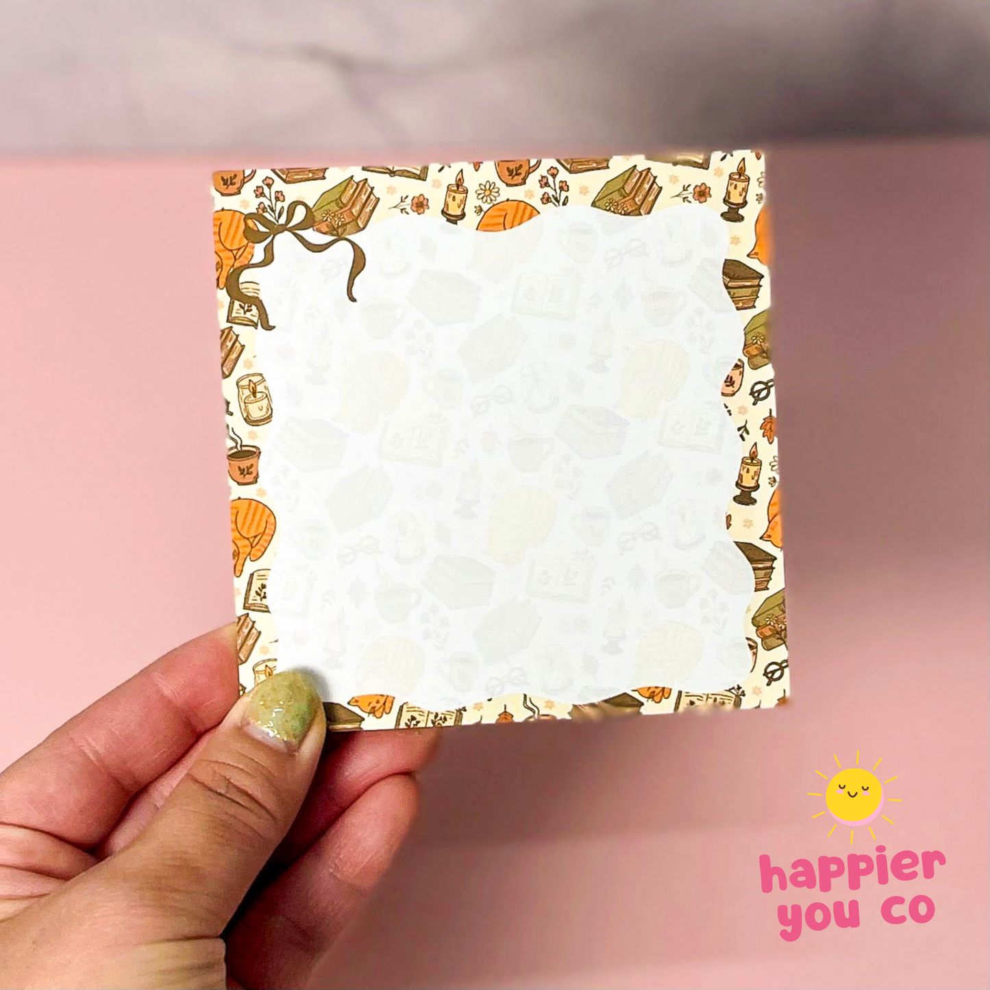 A hand holding a square notepad with a cute illustrated border of cupcakes, coffee cups, and pastries on a pink background. The notepad has a white center with faint pastel cupcake patterns.