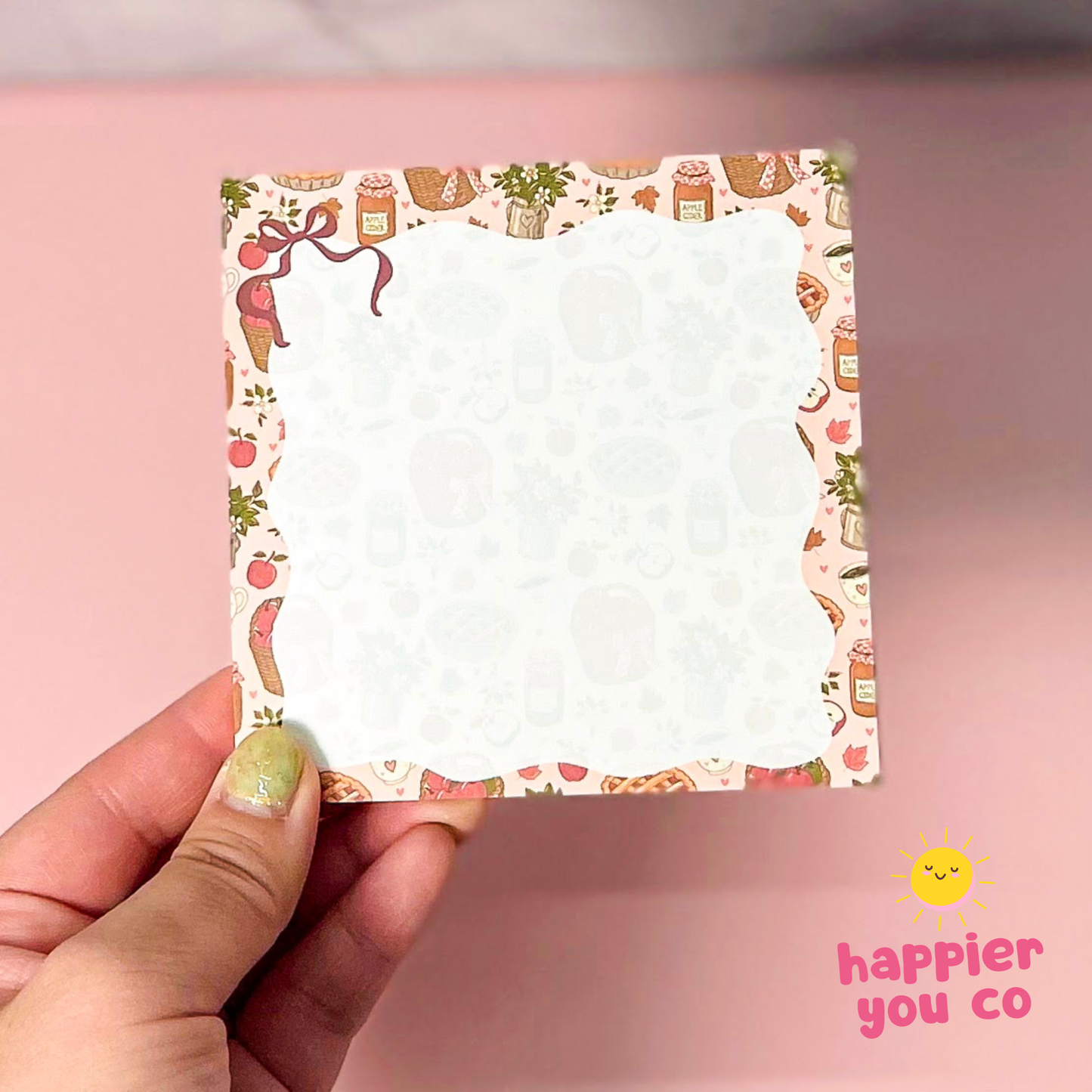 A hand with glittery green nail polish holds a square white notepad with a cute, colorful illustrated border. The background is a soft pink, and a 'happier you co' logo is visible in the bottom right corner.