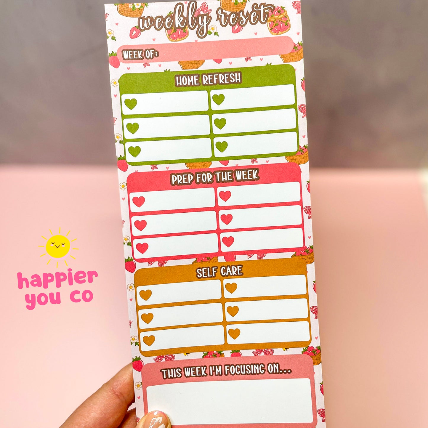 Weekly reset planner with colorful strawberry design held by a hand, featuring 'happier you co' branding.