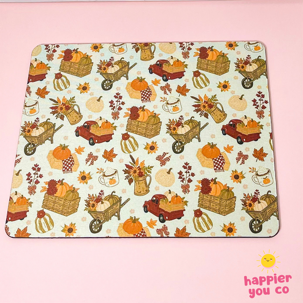 A rectangular mousepad with a light cream background and an autumn harvest pattern featuring pumpkins, sunflowers, leaves, and carts, placed on a solid pink background. A 'happier you co' logo is visible in the bottom right corner.