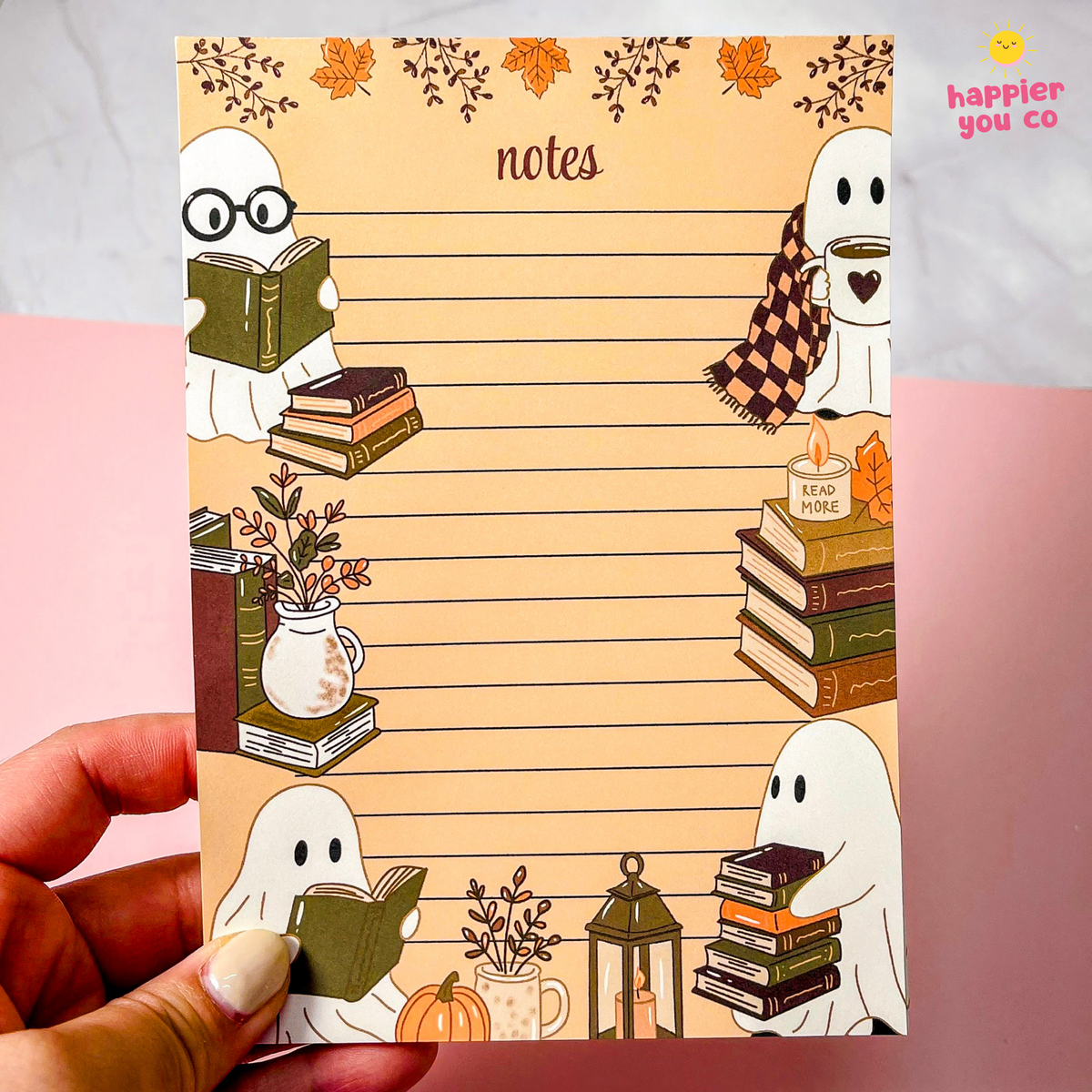 A hand holds a cute autumn-themed notepad with lined writing space and illustrations of ghosts, books, pumpkins, and fall leaves. The notepad has a warm beige background with orange and brown accents.