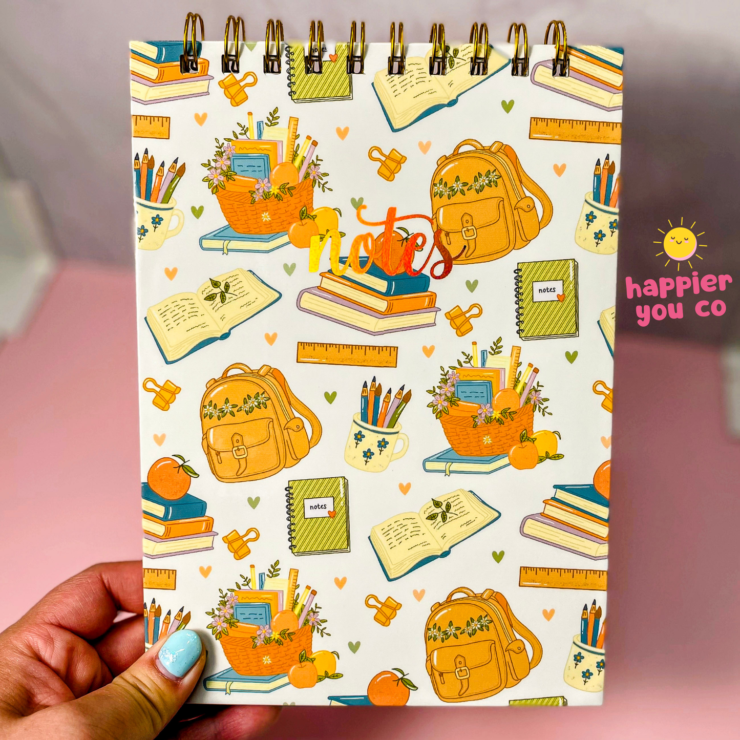 A spiral-bound notepad held by a hand, featuring a colorful school supplies pattern with backpacks, books, pencils, and a heart-shaped 'notes' design. The background is a soft pink surface.
