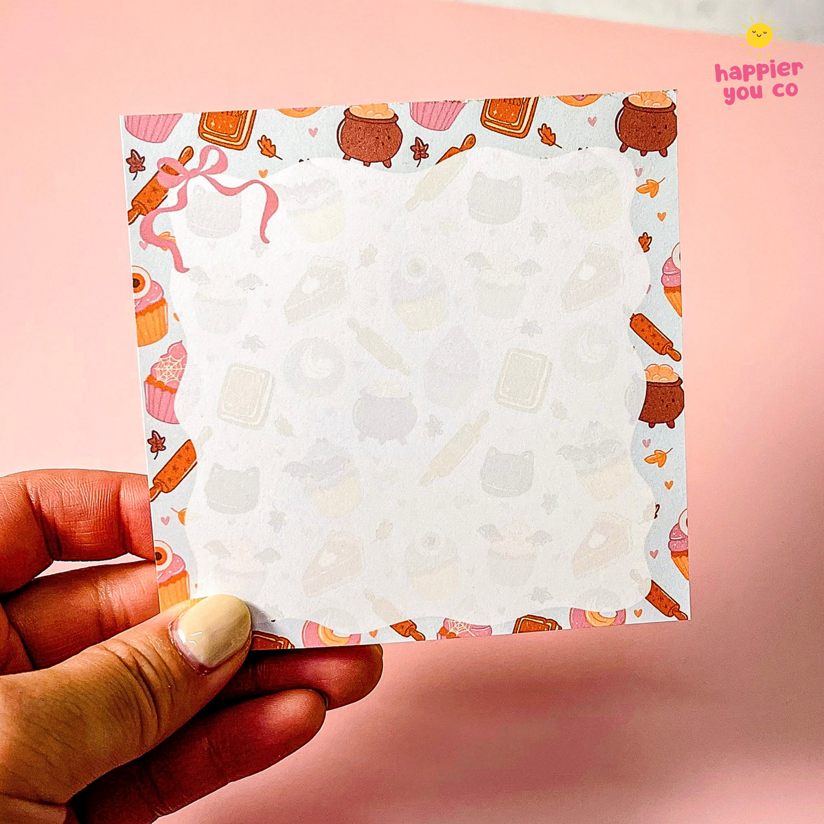A hand holding a square white notepad with a cute pastel patterned border featuring cupcakes, cookies, and small illustrations, against a soft pink background. The brand name 'happier you co' is visible in the top right corner.