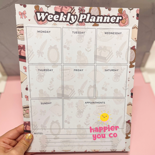 Weekly planner with cartoon designs and 'happier you co' branding held by a hand.