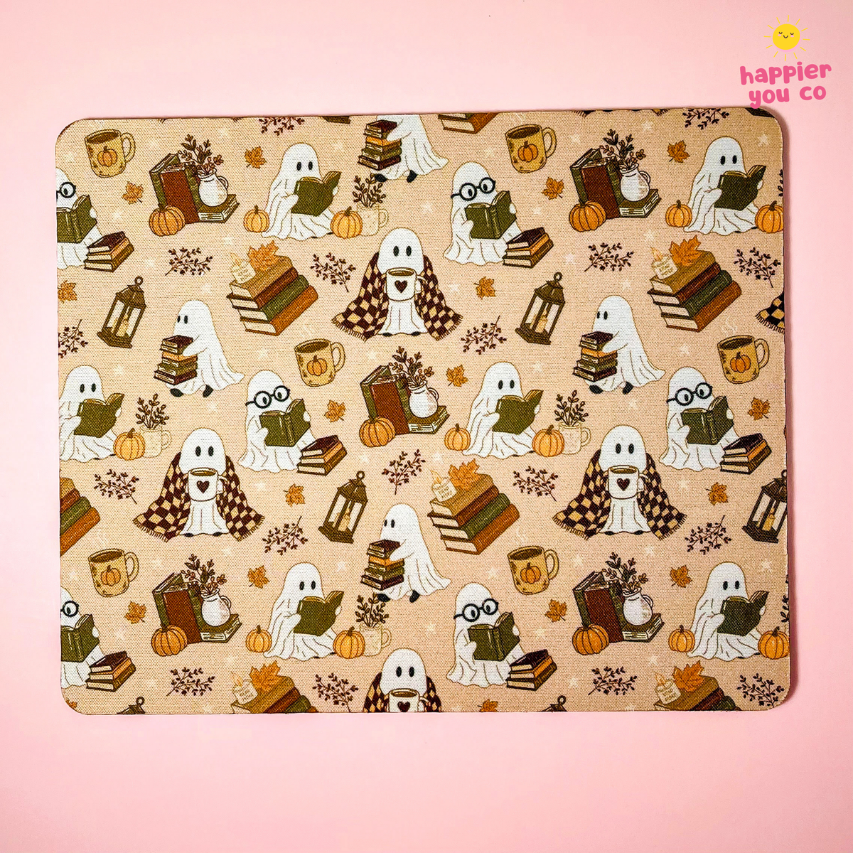 A rectangular mousepad with rounded corners featuring a repeating Halloween pattern of ghosts reading books, pumpkins, lanterns, and autumn leaves on a beige background, set against a solid pink backdrop. A 'happier you co' logo is visible in the top right corner.