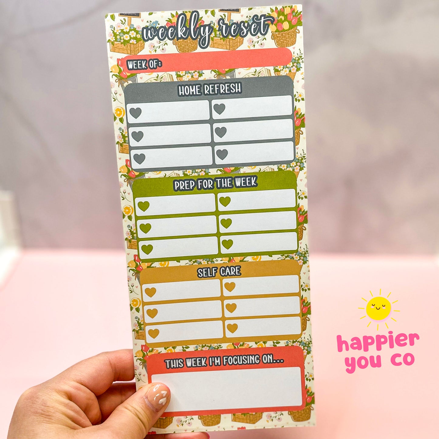 Weekly reset planner pad held by a hand with 'happier you co' branding with flower background