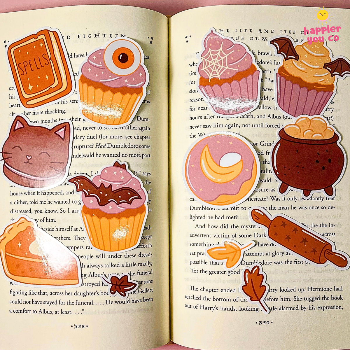 A set of cute, illustrated cupcake-shaped stickers arranged on the pages of an open book. The stickers feature various colorful designs including pink, orange, brown, and yellow frosting, with whimsical details like spiderwebs, candy canes, and chocolate drizzle.