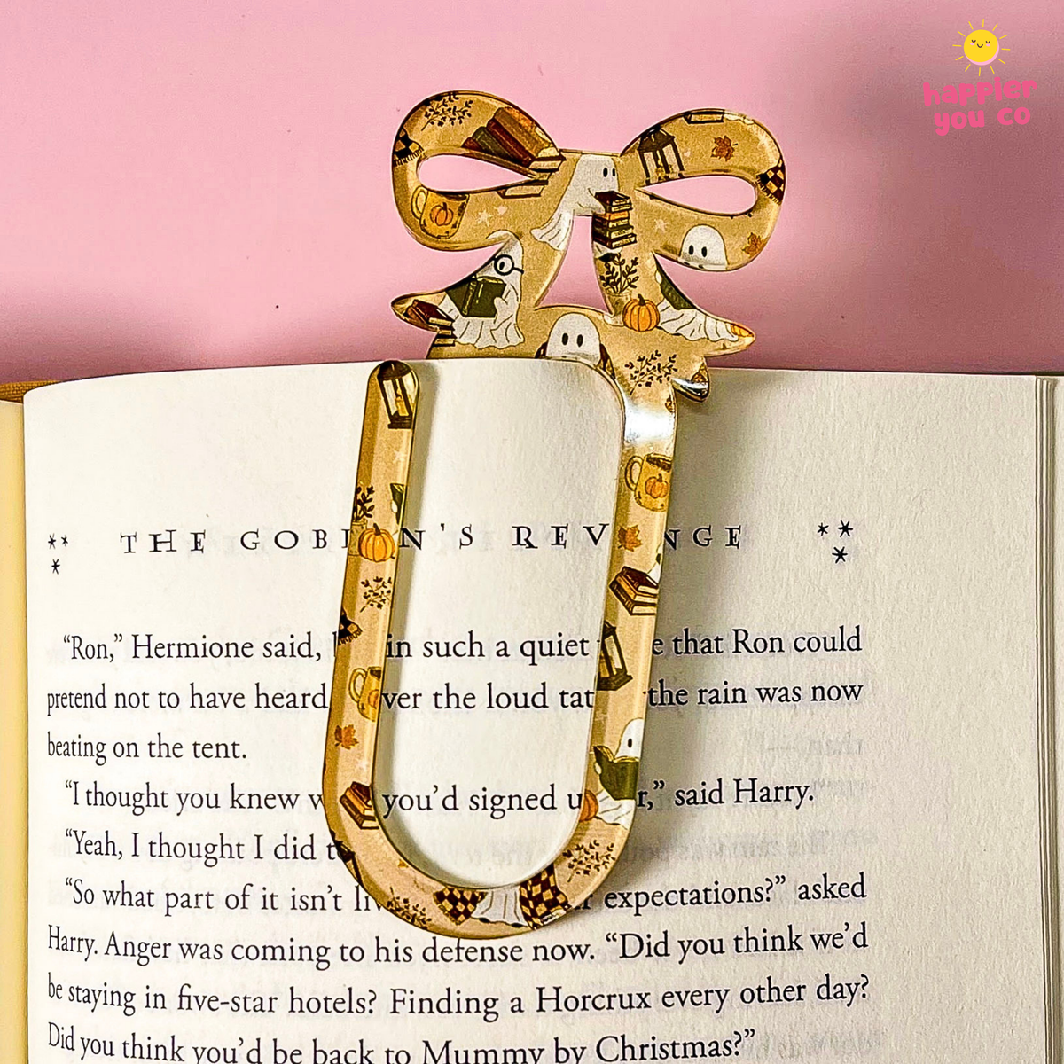 A Halloween-themed metal bookmark shaped like a bow, featuring a pattern of ghosts, pumpkins, and spiderwebs on a gold background, held open in an open book against a pink background.