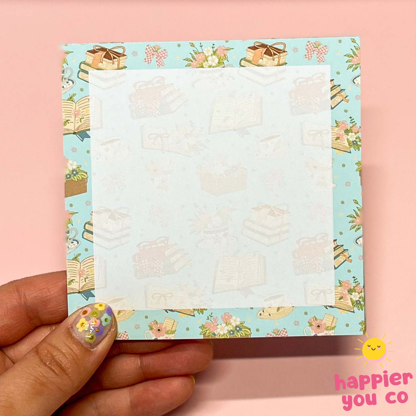 Hand holding a small notepad with a floral and book design on a pink background