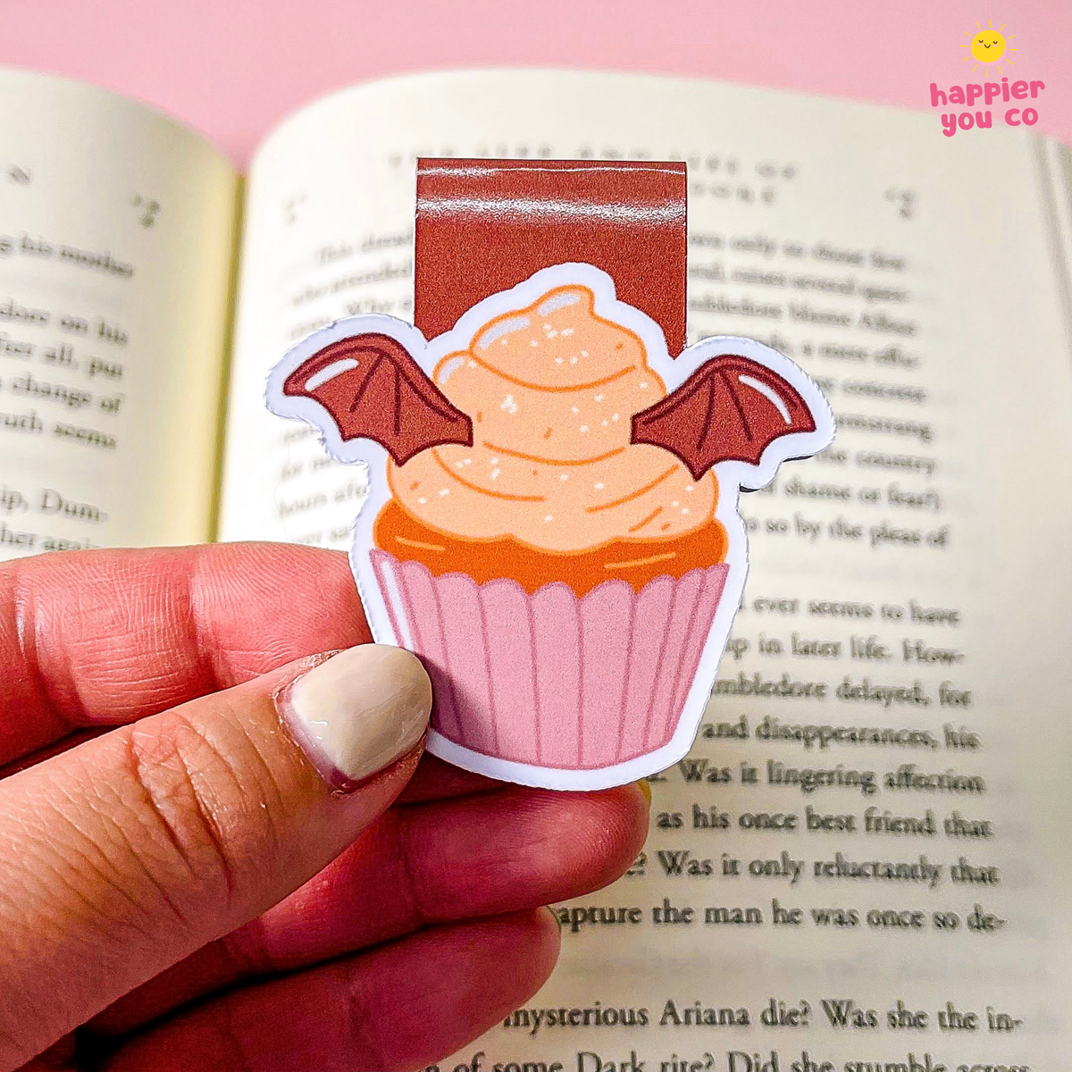 A hand holds a die-cut bookmark shaped like a pink cupcake with orange frosting and brown bat wings, positioned over an open book. The sticker has a brown rectangular tab at the top.