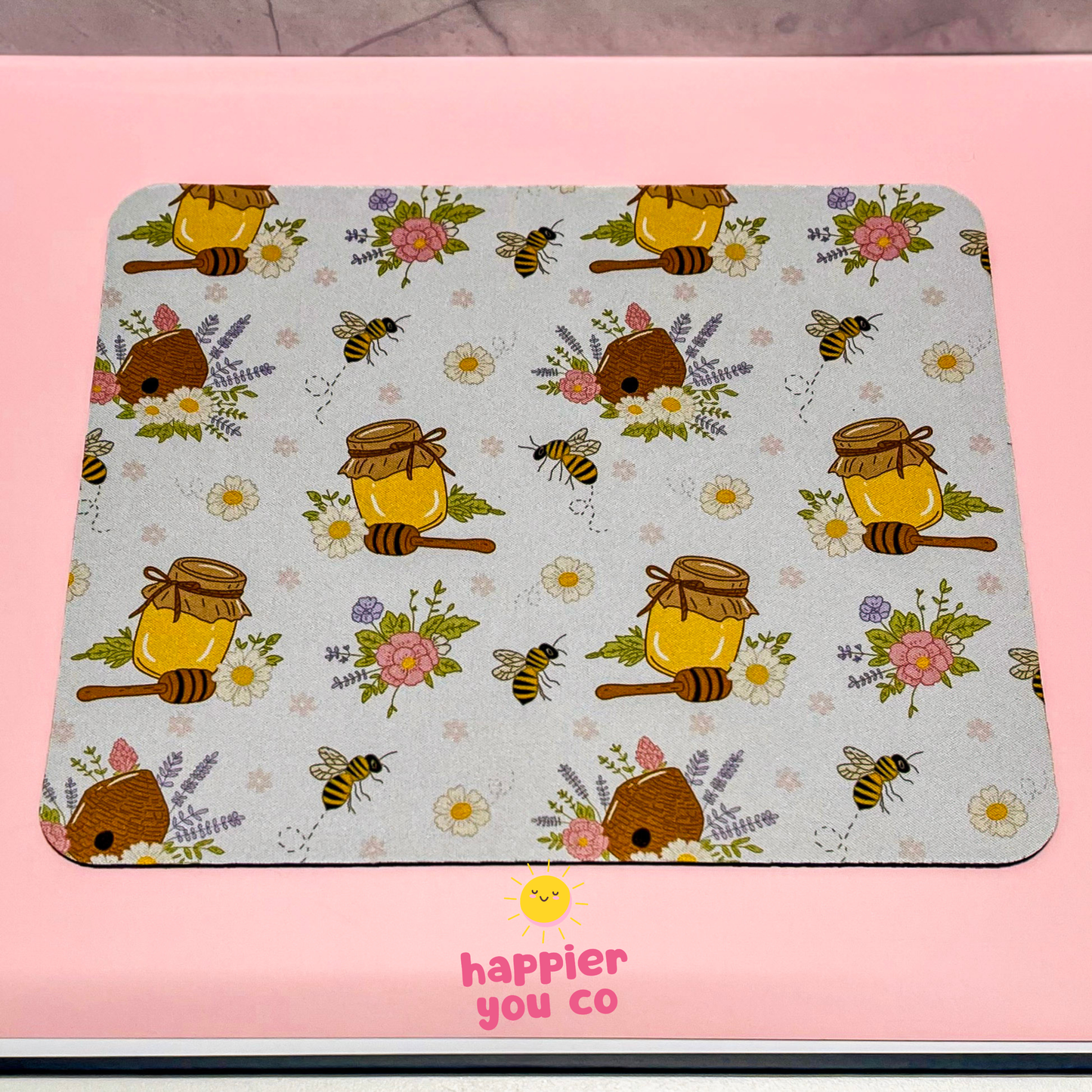 Mouse pad with honeycomb, bees, and flowers pattern on a pink surface with 'happier you co' branding.