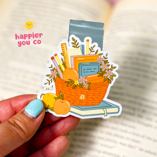 A hand with light blue nail polish holds a die-cut sticker featuring a wicker basket filled with books, pencils, flowers, and fruit, set against an out-of-focus open book background. Pink text in the upper left reads 'happier you co'.