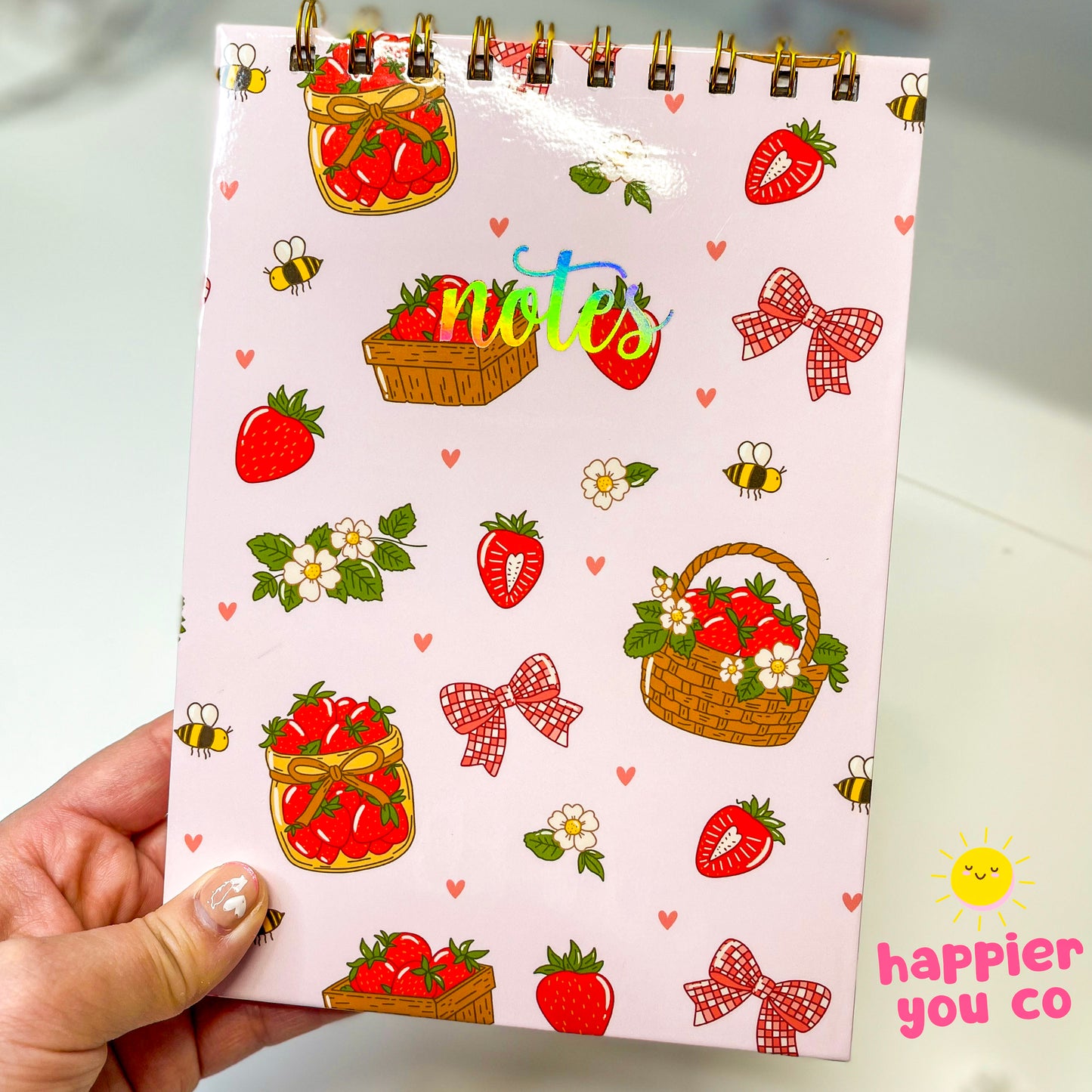 Notebook with strawberry and basket design held by a hand, featuring 'happier you co' branding.