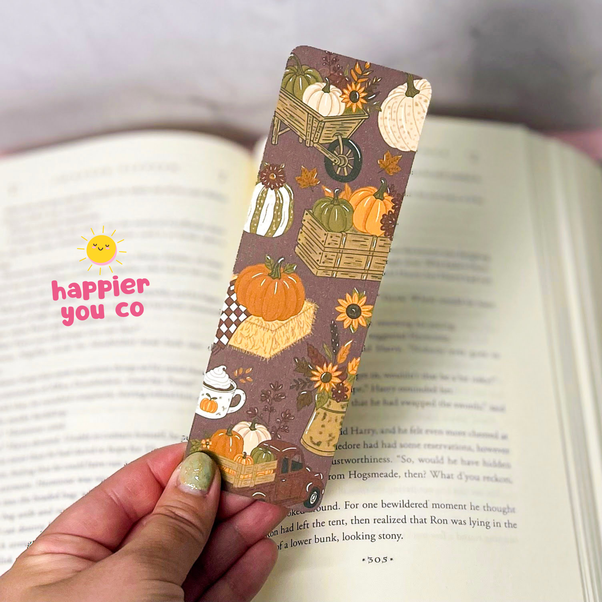 A hand holds a brown autumn-themed bookmark with a pumpkin, acorn, sunflower, and basket pattern over an open book. A pink 'happier you co' logo is visible on the left side of the image.