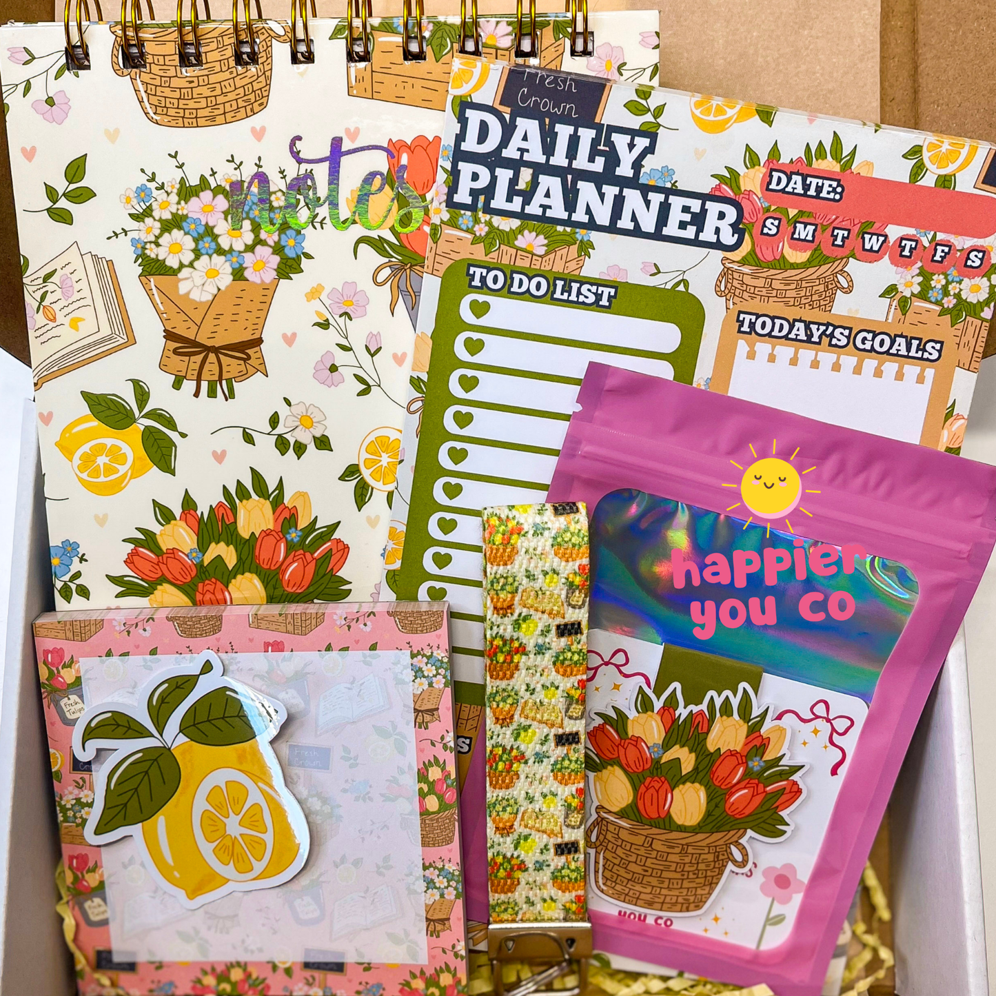 Collection of colorful planners and a bag with 'Happier You Co' branding.