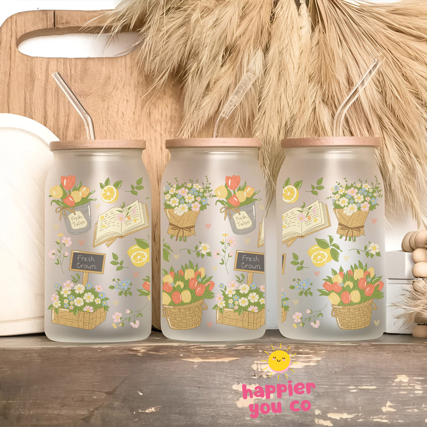 Three glass jars with floral designs and straws on a wooden surface, featuring the brand 'happier you co'.