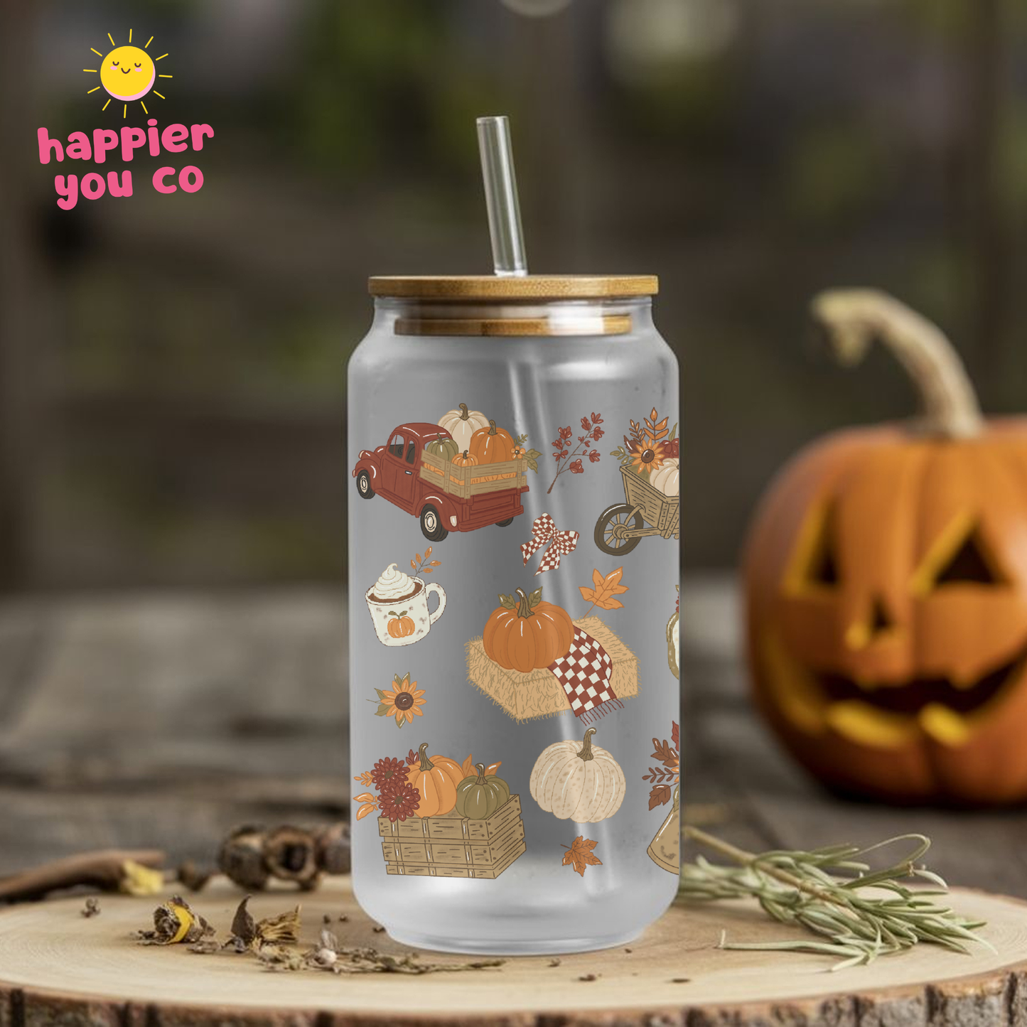 A frosted glass can-style tumbler with a bamboo lid and clear straw, decorated with autumn-themed illustrations including pumpkins, mugs, autumn leaves, pumpkin truck. The tumbler is shown outdoors on a wooden surface with a blurred pumpkin and foliage in the background.
