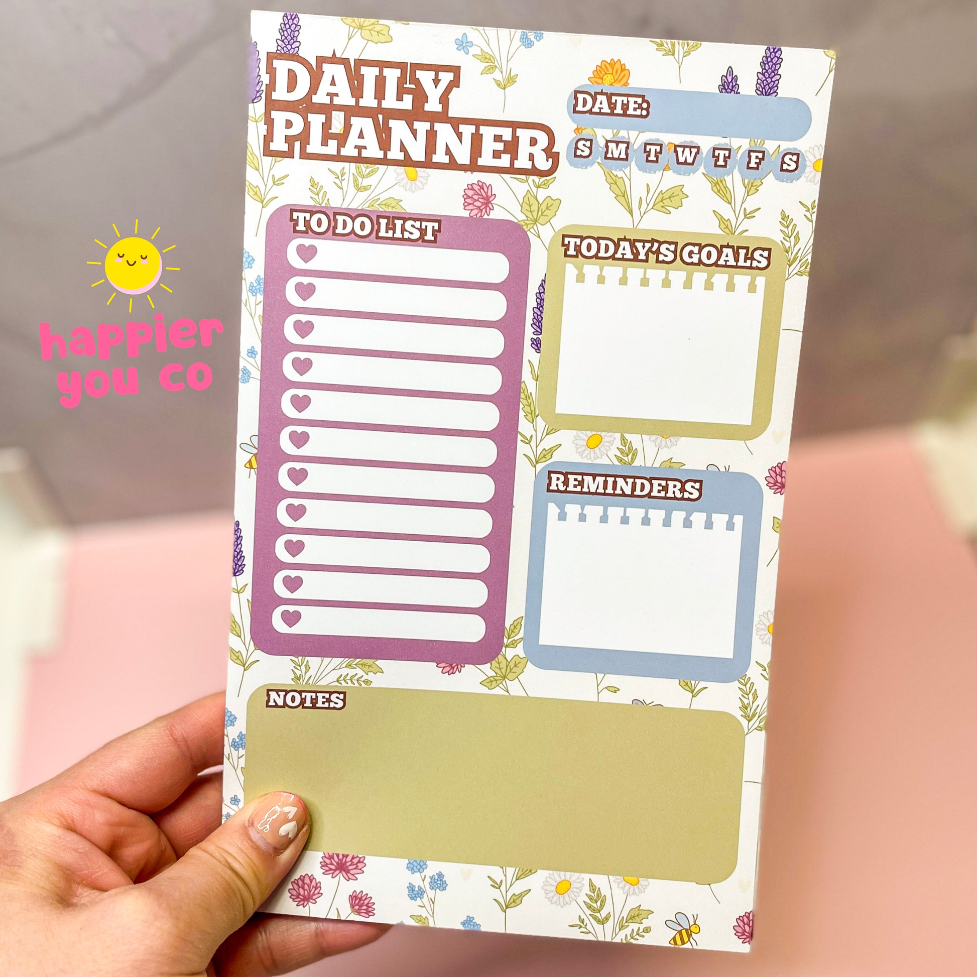 Daily planner with floral design held by a hand, featuring sections for to-do list, goals, reminders, and notes.