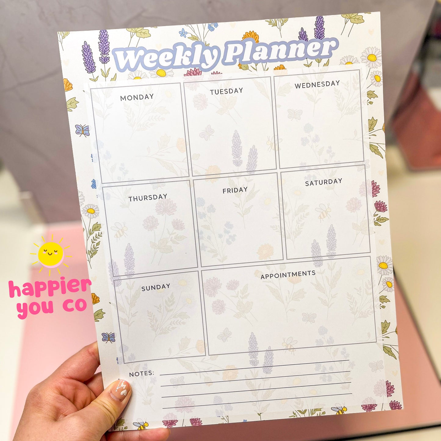 Weekly planner with floral design held by a hand, branded 'happier you co'.