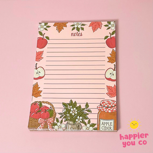 A pink lined notepad with an autumn-themed border featuring apples, leaves, flowers, and an apple cider jar, photographed on a solid pink background. A small 'happier you co' logo appears in the bottom right corner.