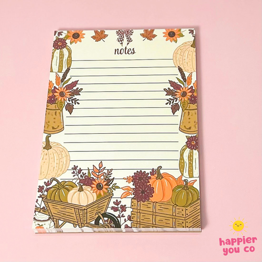 An autumn-themed lined notepad with a floral border featuring pumpkins, sunflowers, and fall leaves, displayed on a solid pink background.