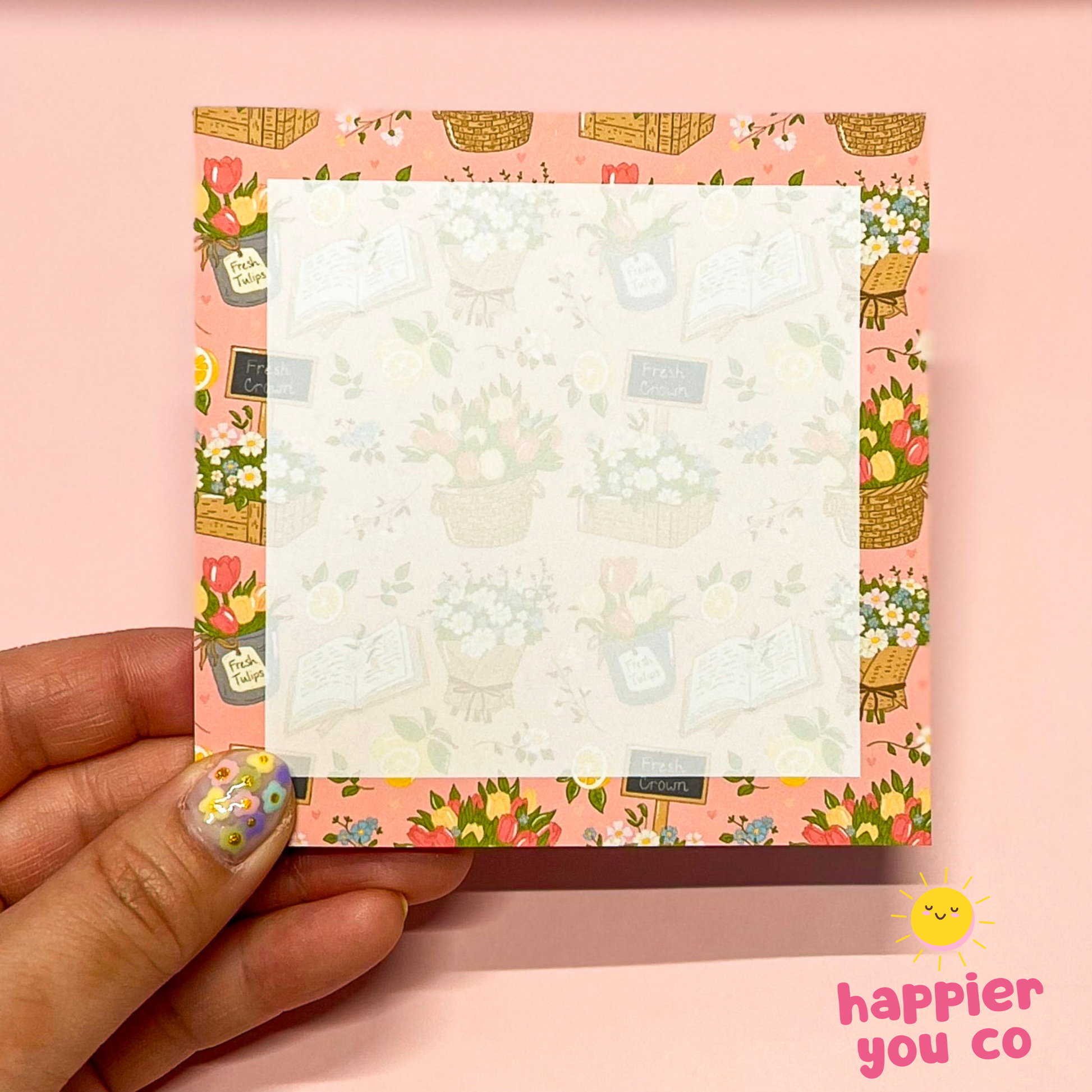 Floral-patterned notepad held by a hand on a pink background with 'happier you co' branding.