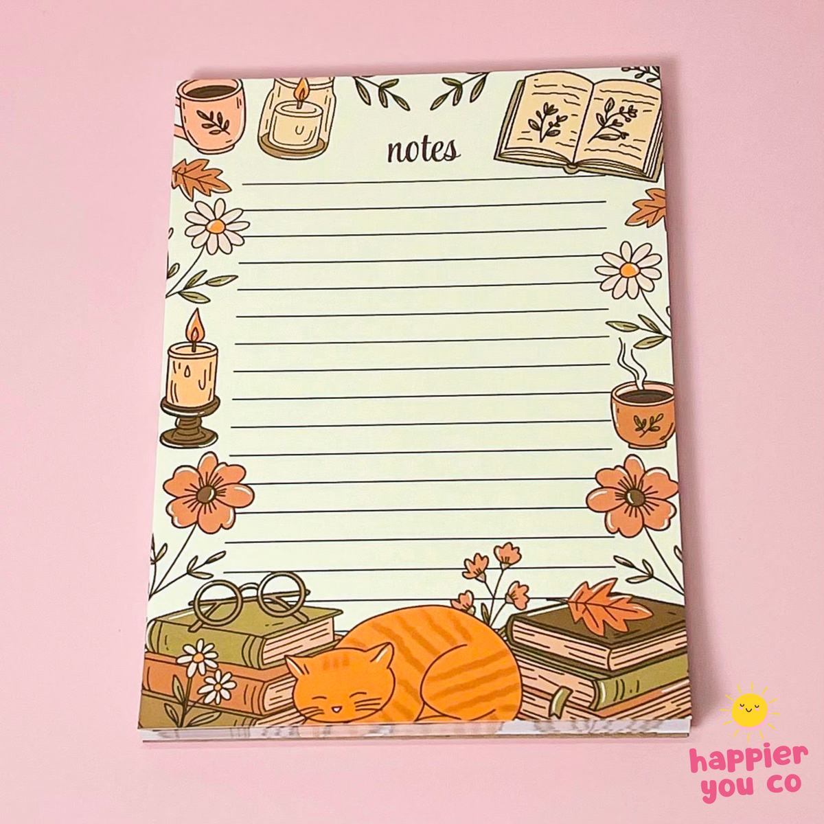 A lined notepad with a decorative autumn-themed border, featuring illustrations of candles, flowers, books, glasses, and a sleeping orange cat, photographed on a pink background. The notepad is branded with 'happier you co' in the bottom right corner.