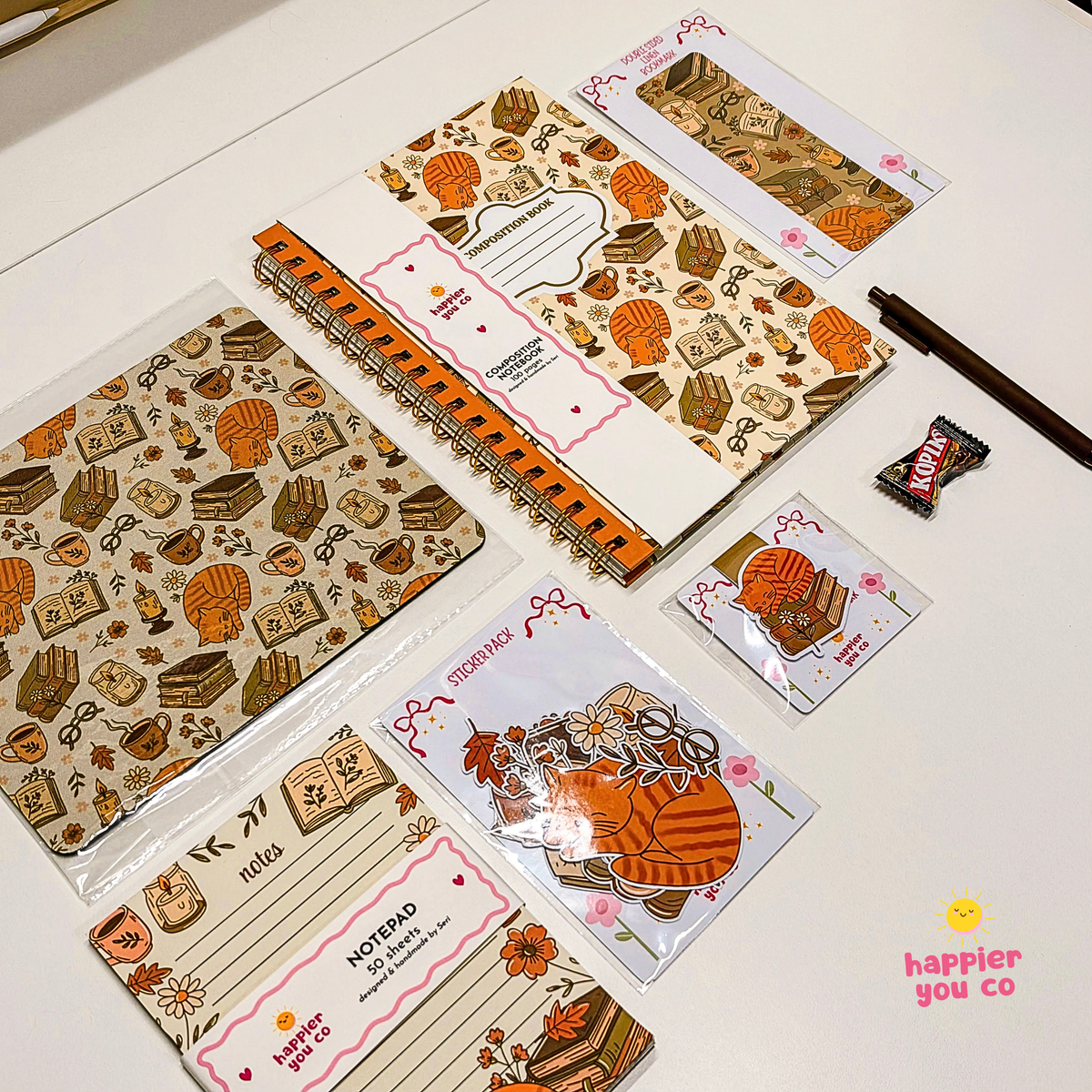 A coordinated stationery set arranged on a white surface, featuring patterned notebooks, cards, stickers, and a pen with autumn-themed illustrations such as pumpkins, books, and flowers. A small candy wrapper is also visible among the items.