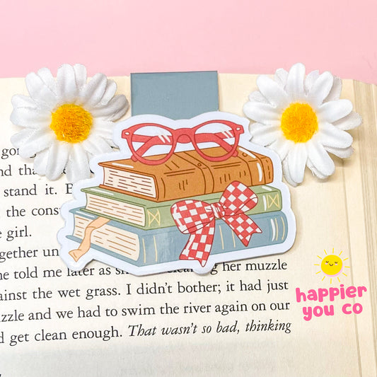 Illustration of books with a bow and sunglasses on a page with flowers, featuring 'happier you co' branding.