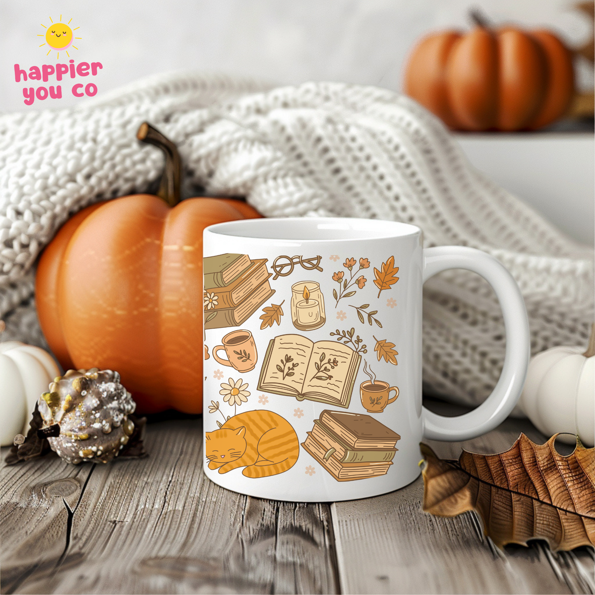 A white ceramic mug with a cozy autumn-themed illustration of books, coffee cups, leaves, and a sleeping cat, styled on a wooden surface with pumpkins, a knit blanket, and fall leaves. The happier you co logo is visible in the top left corner.