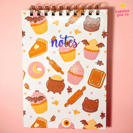 A spiral-bound notebook with a white cover decorated with cute autumn-themed illustrations of cupcakes, donuts, pies, cauldrons, and leaves, set against a solid pink background.