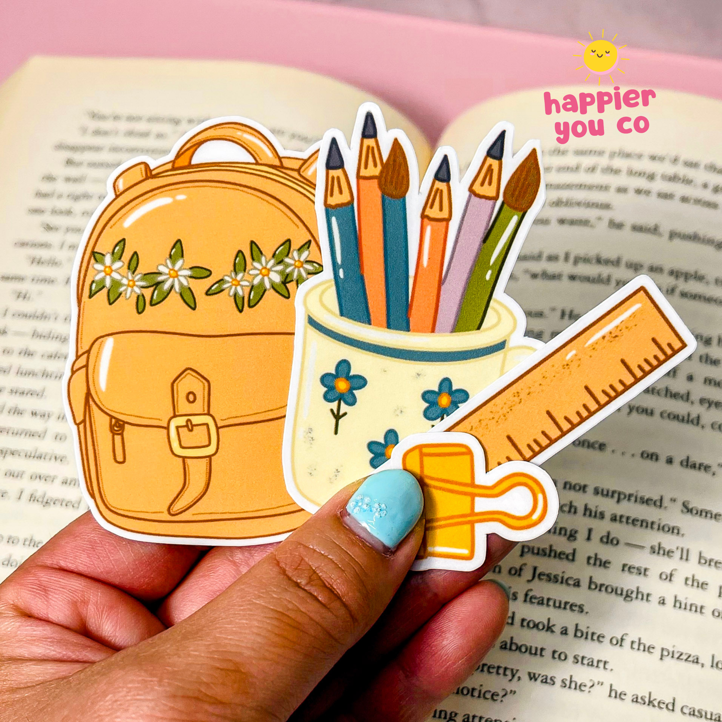 A hand holds a die-cut sticker featuring a school backpack, pencils, a cup, and a ruler over an open book. The sticker has a white border and is shown with a pink background and a 'happier you co' logo in the top right corner.
