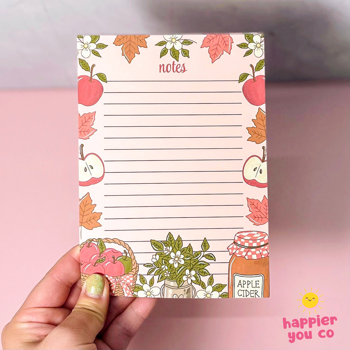 A hand holding a square notepad with an autumn apple theme, featuring lined writing space and illustrations of apples, leaves, flowers, and an apple cider jar, set against a pink background.