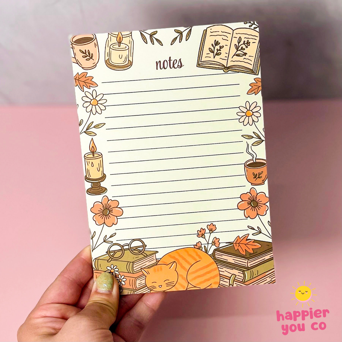 A hand holds a cream-colored lined notepad decorated with autumn-themed illustrations, including candles, flowers, books, and a sleeping cat, against a pink and gray background.