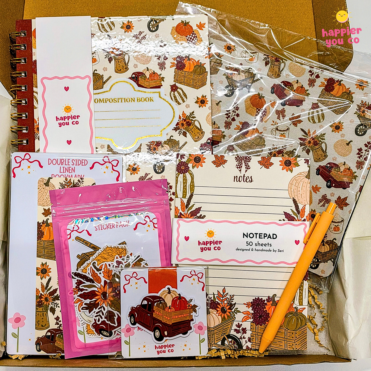 A stationery gift set arranged in a box, featuring multiple printed pads, stickers, and a notepad with autumn-themed illustrations such as pumpkins, fall leaves, cactus, and trucks. Visible text includes 'happier you co' and product labels like 'Composition Book,' 'notes,' 'notepad 50 sheets,' and 'double sided linen.