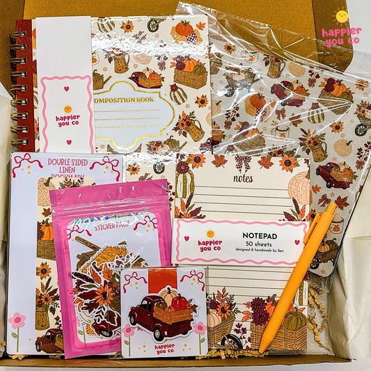 A stationery gift set arranged in a box, featuring multiple printed pads, stickers, and a notepad with autumn-themed illustrations such as pumpkins, fall leaves, cactus, and trucks. Visible text includes 'happier you co' and product labels like 'Composition Book,' 'notes,' 'notepad 50 sheets,' and 'double sided linen.