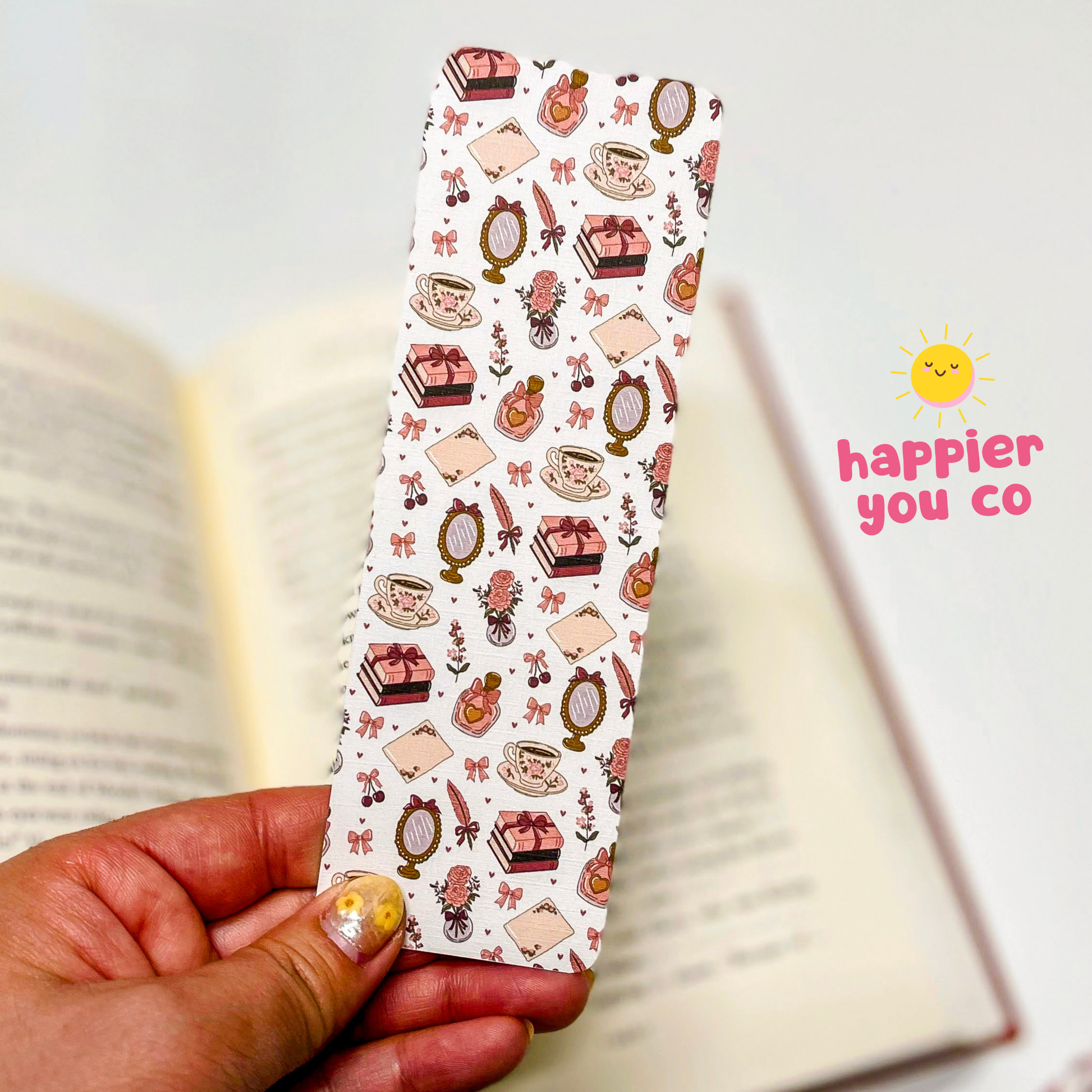 Decorative bookmark with illustrations held over an open book, featuring the brand 'happier you co'.