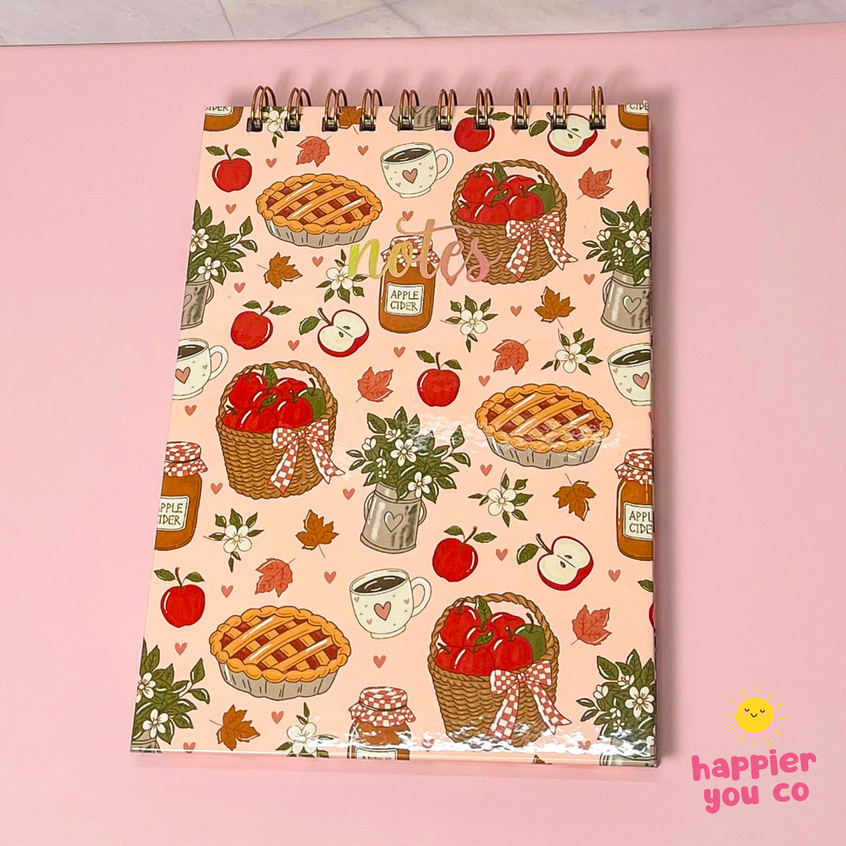 A spiral-bound notebook with a warm autumn apple-themed pattern, featuring illustrations of pies, baskets of apples, jars of apple pie sauce, and hearts on a light pink cover, displayed on a solid pink background.