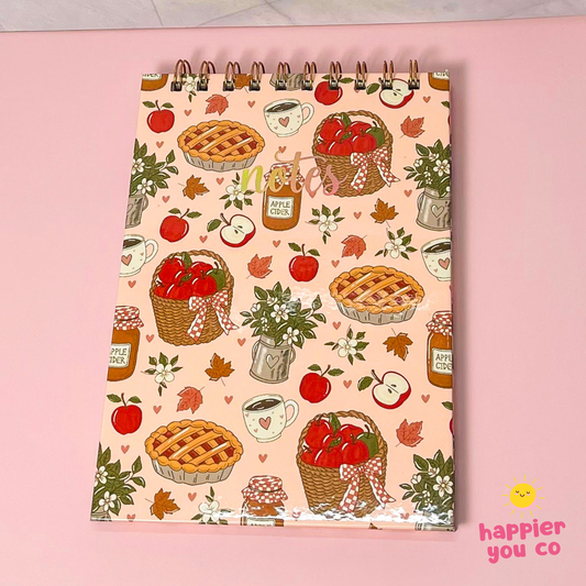 A spiral-bound notebook with a warm autumn apple-themed pattern, featuring illustrations of pies, baskets of apples, jars of apple pie sauce, and hearts on a light pink cover, displayed on a solid pink background.